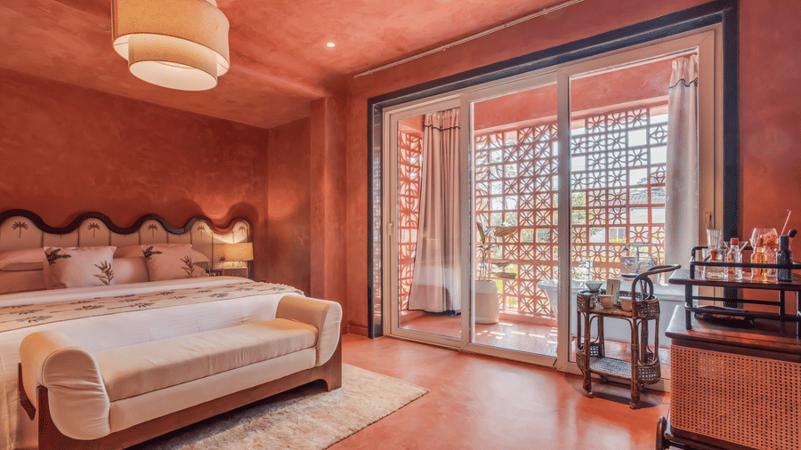 Bedroom at Unseen Boutique Hotel with reddish?brown walls, tufted headboard, white rug, sliding glass door to balcony, and side cart with bottles and glasses.