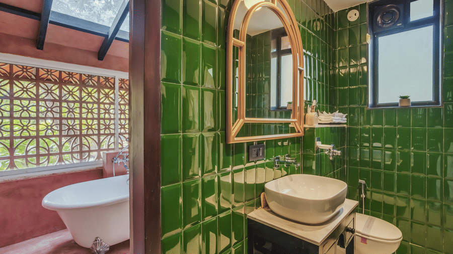 Bathroom at Unseen Boutique Hotel with green glossy tiled walls, arched wooden mirror, ceramic sink, and adjoining claw?foot bathtub.