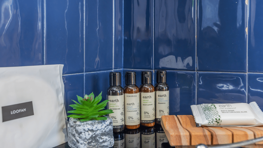 Bathroom counter at Unseen Boutique Hotel with toiletries, folded towel, potted plant, bottles, and white box on blue tiled surface.