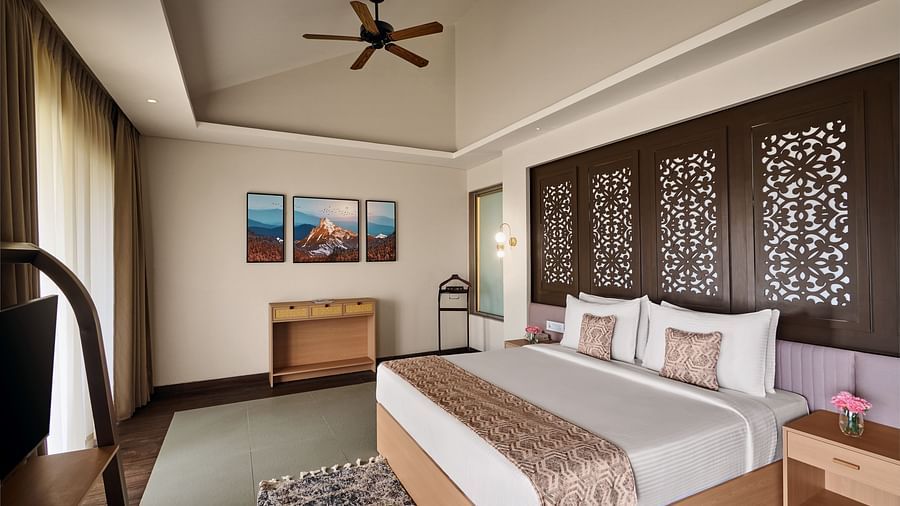 Stylish hotel bedroom with a large bed, decorative wooden panel headboard, ceiling fan, framed artwork on the wall, and floor-to-ceiling curtains - c