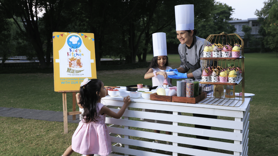 An outdoor food counter with 2 chefs where a child chef and a woman chef are standing behind a live cooking setup at The Golden Tusk, Jim Corbett.