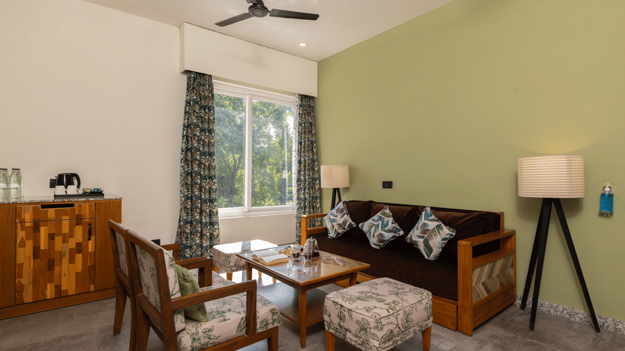 Living area with wooden furniture, including a sofa and chairs, a floor lamp, and a view of greenery outside at The Golden Tusk, Jim Corbett