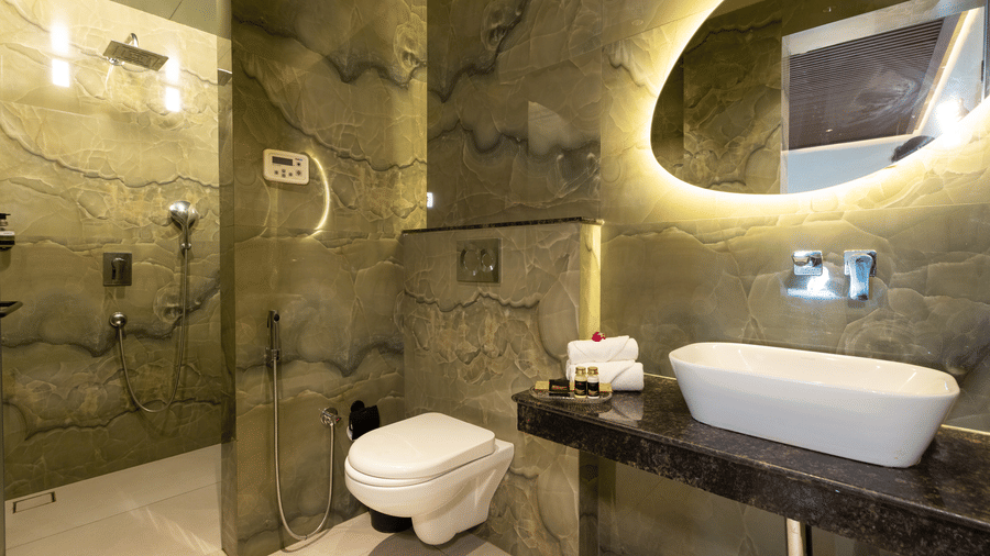 A contemporary bathroom with a wall-mounted toilet, a sink on a dark counter, a circular backlit mirror, and a walk-in shower area at The Golden Tusk, Jim Corbett