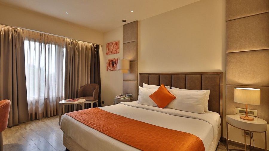 A comfortable hotel room with a bed, a small seating area by the window, and decorative art on the wall above the headboard. (Hotel Saket 27, New Delhi).