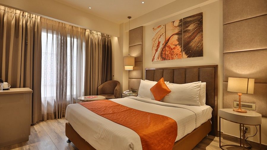A hotel room with a neatly made bed with orange and white bedding, a window with sheer and blackout curtains, a small seating area, and abstract artwork. (Hotel Saket 27, New Delhi).
