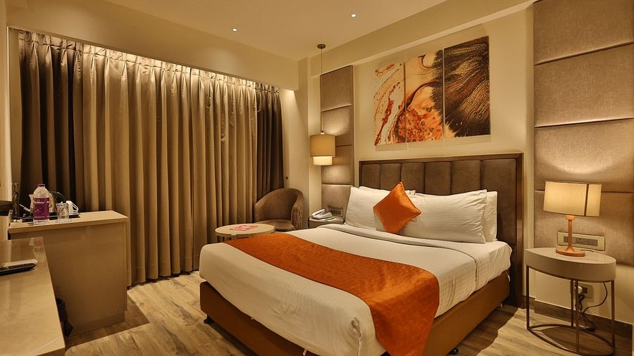 A wide shot of a contemporary hotel room with a large bed featuring white and orange bedding, a bedside table with a lamp, a small seating area, and curtains covering a window. (Hotel Saket 27, New Delhi).