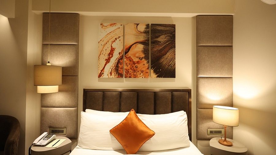 A well-lit hotel room with a large bed, abstract art above the headboard, and two side tables with lamps. (Hotel Saket 27, New Delhi)