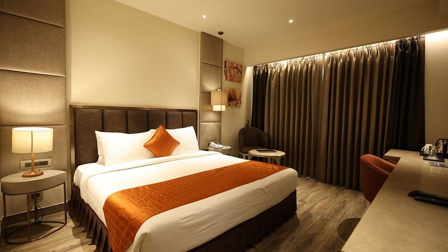 A wide shot of a contemporary hotel room showing a bed with orange and white linens, bedside tables with lamps, a seating area, and a desk with a chair. (Hotel Saket 27, New Delhi).