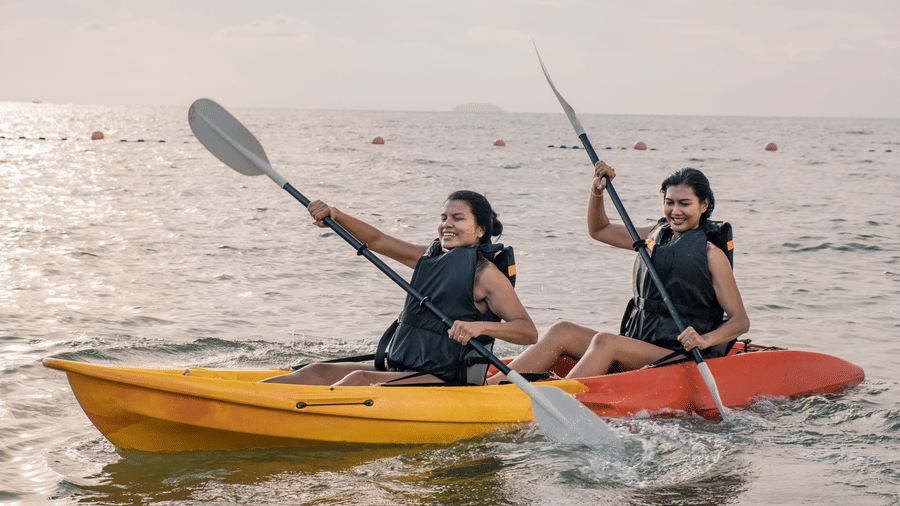2 women in separate kayaks on the sea, paddling with smiles, enjoying a beautiful sunset, reflecting a calm and joyful water activity.