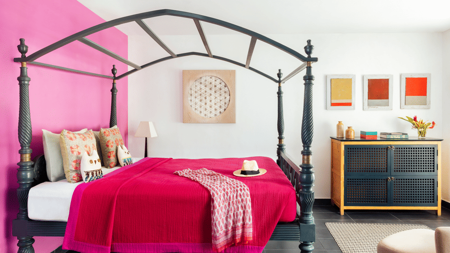A view of a bed at Owl and the Pussycat Hotel in Galle with a high canopy frame siting against a vibrant wall, adjacent to a chest of drawers with framed art and floral arrangements.