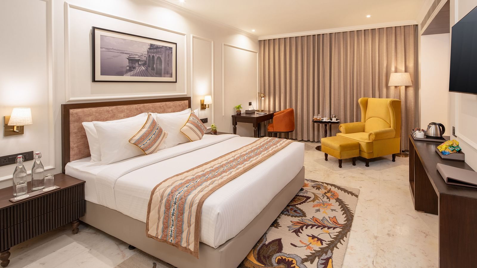 An image of a bedroom with bedside table, photo frame hanged on the wall, work desk with sofa chair, and a flat TV screen - Brij Eternity, Vrindavan