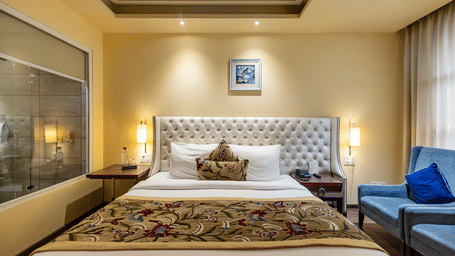 A bedroom with a king size bed, side tables, lamps, sofa, and a glass partition visible at Indraprastha Resort & Spa, McLeod Ganj.