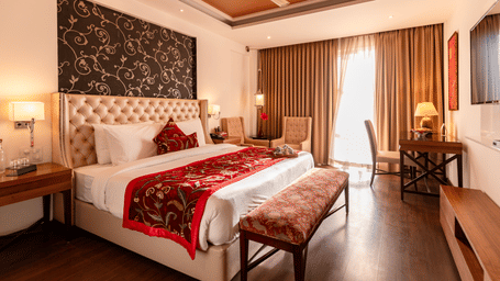 A bedroom with a king size bed, side tables, lamps, bench, curtains, and wall-mounted TV inside at Indraprastha Resort & Spa, McLeod Ganj.