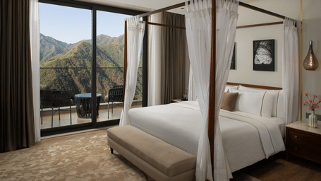 Bedroom with white four-poster canopy bed, bench, and sliding glass balcony doors looking out to mountains.