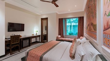 A headboard view of a spacious bedroom at Panna Vilas Palace, displaying a wall-mounted television with a wooden table underneath and large windows inviting natural light