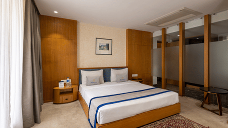 Wide view of the bedroom of Suite at Pravasa Stay Vijay Nagar, Indore, showcasing its queen size bed, bedside tables, and tinted glass partitions.