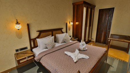 A cosy bedroom featuring a wooden bed frame, warm wall-mounted lamps, and a decorative towel arrangement - Tathastu Chavri Resort, Jawai