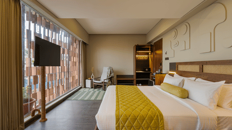 A modern hotel room with a large bed featuring white linens and a mustard?yellow quilted throw, floor?to?ceiling window with sheer curtains, mounted flat?screen television, lounge chair near the window, desk area with shelving, and walls decorated with rec