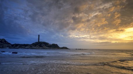 A farout view of kega lighthouse in the distance with waves overlapping the beach and sun setting in the background.