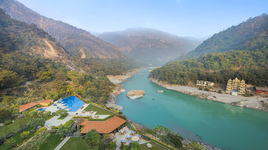 Riverside patio dining area overlooking a turquoise river, mountain landscape, landscaped lawns, and an outdoor pool beside the hotel.
