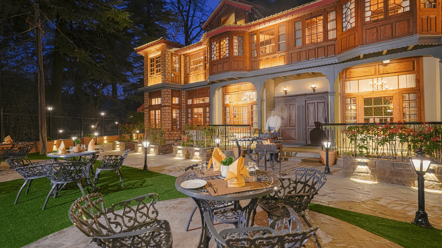 The facade of BluSalzz Terrah Hills Resort, Dalhousie, featuring illuminated lights, balconies, windows, and an outdoor seating area with tables, and chairs in the foreground at night .