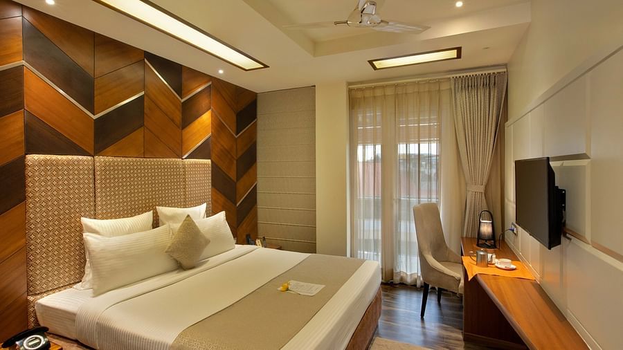 Image of warmly lit Coral Suite at Coral Reef Hotel and Spa in Port Blair featuring a plush bed in front of a TV mounted on the wall and next to it is a work desk and a chair near the door to balcony