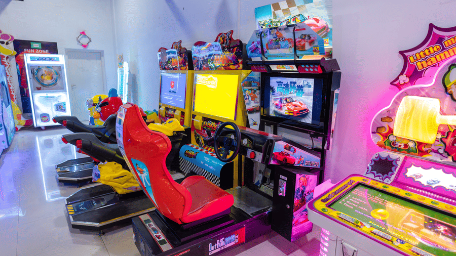 Racing simulator machine at the DSF Fun Zone, DSF Grand Plaza, Tuticorin, with a red bucket seat, steering wheel, and screen showing a racing game, beside motorbike ride games.
