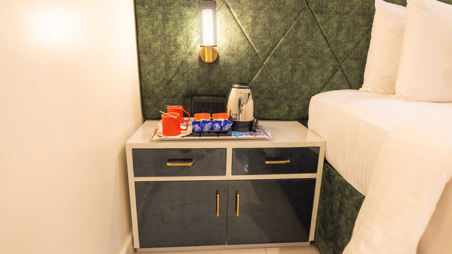 Bedside table at DSF Grand Plaza, Tuticorin with a tea and coffee tray, a kettle, red mugs, a wall lamp, and a green quilted headboard beside the bed.