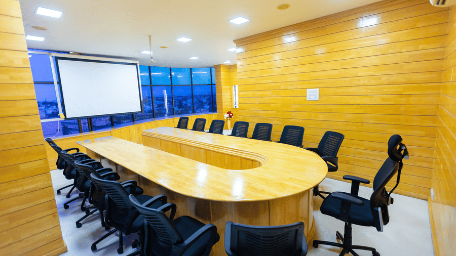 The business centre at DSF Grand Plaza, Tuticorin, with a U-shaped wooden conference table, black mesh chairs, and a projector screen beside floor-to-ceiling windows.