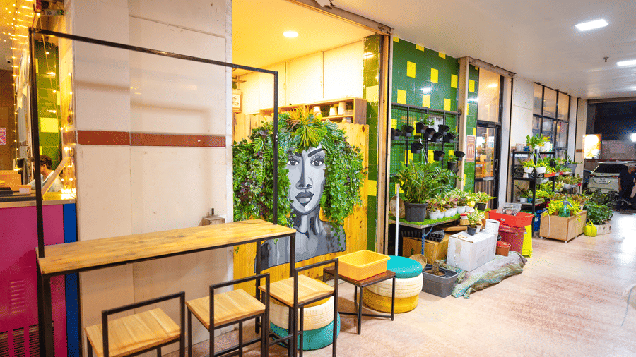 Small cafe seating area beside a plant nursery, with a leafy mural of a face on the wall, high wooden tables, and tyre-stool seating.