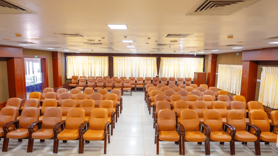 Conference hall at DSF Grand Plaza, Tuticorin viewed from the stage, showing rows of orange chairs split by a centre aisle and cream curtains.