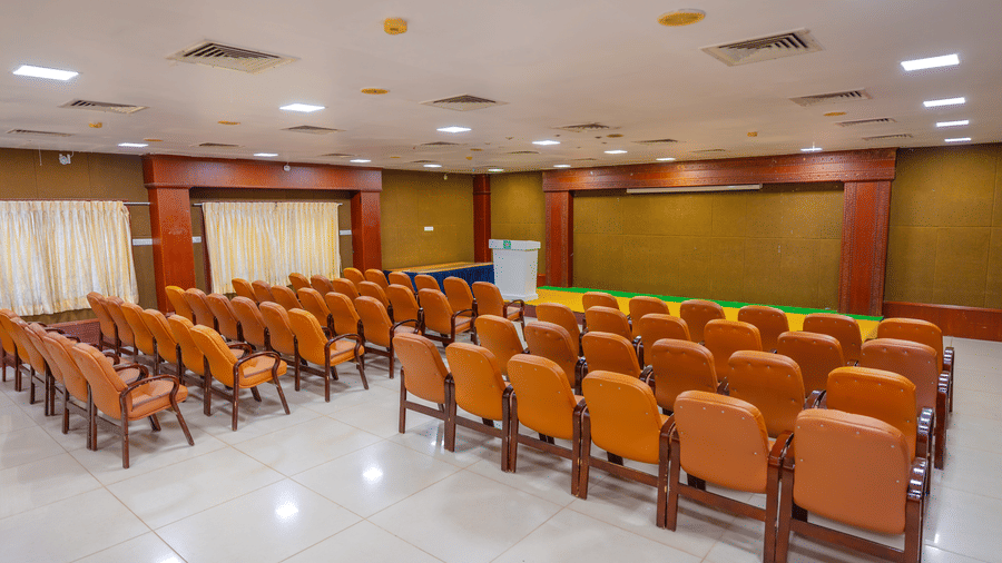 Conference hall at DSF Grand Plaza, Tuticorin with curved rows of orange chairs, a raised stage with a green and yellow carpet, and cream curtains.