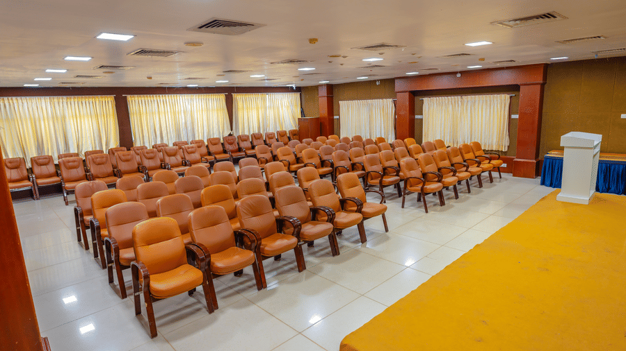 Conference hall at DSF Grand Plaza, Tuticorin with rows of orange chairs facing a stage with a yellow carpet, cream curtains, and a corner lectern.