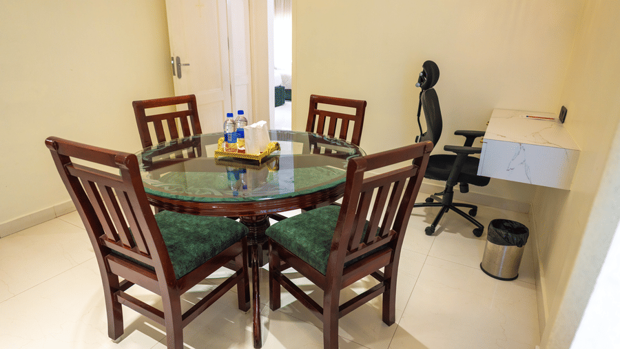 Suite dining nook at DSF Grand Plaza, Tuticorin with a round glass-top table, 4 green chairs, a desk with office chair, and an open door to the bedroom.