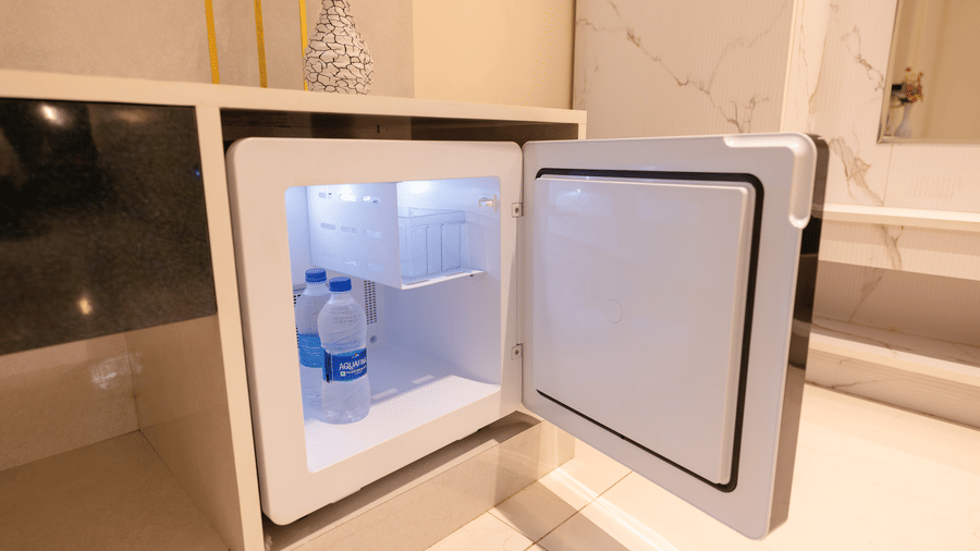 Open minibar fridge in a guest room at DSF Grand Plaza, Tuticorin with 2 Aquafina water bottles inside, set into a cabinet below a marble-clad wall and a decorative vase.