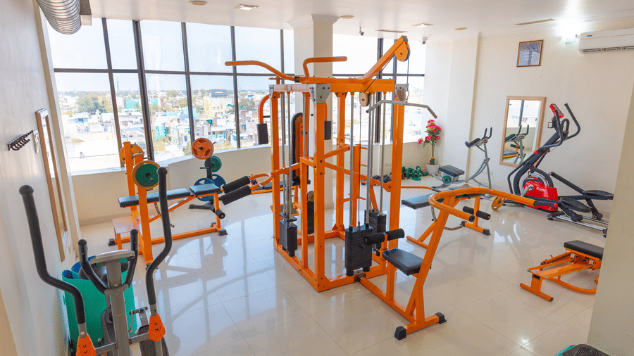 Hotel gym at DSF Grand Plaza, Tuticorin with an orange multi-station cable machine, cross-trainers, a weight bench, and windows overlooking the city.