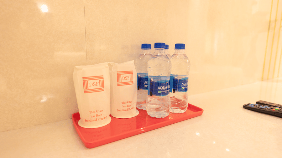In-room amenity tray at DSF Grand Plaza, Tuticorin with 2 DSF-branded sterilised glass covers and 3 sealed Aquafina water bottles on a red tray, with a TV remote alongside.