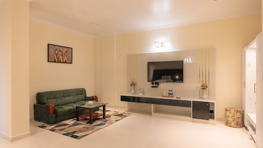 Suite living room at DSF Grand Plaza, Tuticorin with a green sofa, coffee table, a wall-mounted TV console, mini fridge, and a floral ottoman.
