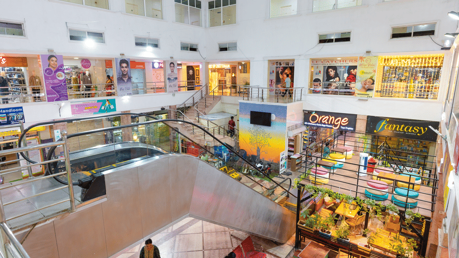 Aerial interior view of a shopping mall atrium with an escalator, multiple storefronts including a gift shop and a music store, a rooftop cafe seating area, and shoppers below.