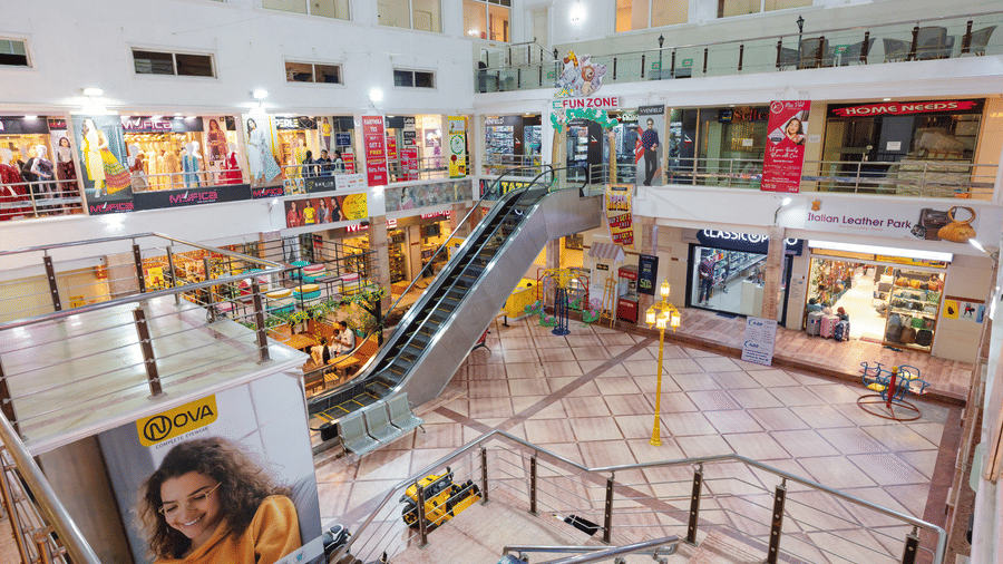 Wide interior view of a shopping mall with an escalator leading to a Fun Zone play area, clothing and leather goods stores, and a carousel ride for children on the ground floor.