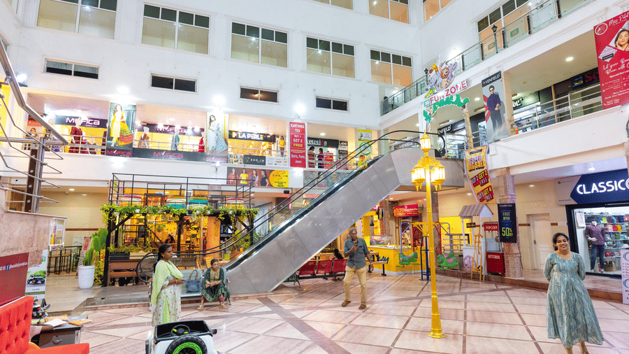 Ground floor of a shopping mall with an escalator, a Fun Zone play area with amusement rides, shoppers walking past storefronts, and a decorative lamp post.
