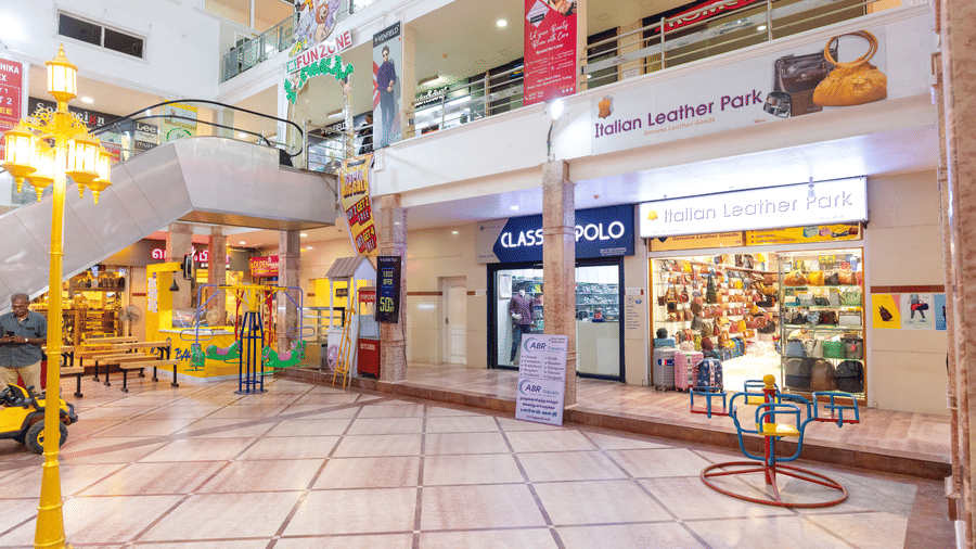 Shopping mall walkway with a leather goods store, a clothing store, an escalator leading to a Fun Zone play area, and a small roundabout ride on the ground floor.