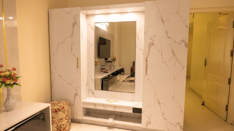 Guest room at DSF Grand Plaza, Tuticorin with a marble-finished wardrobe and mirror unit, backlit ceiling spotlights, a dressing shelf, a floral pouf stool, and a minibar reflected in the mirror.