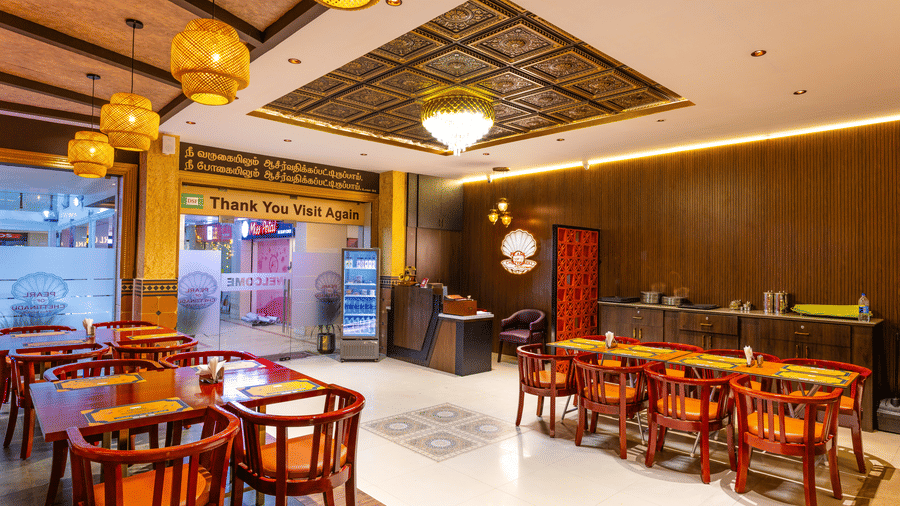 The interior of Pearl of Chettinadu restaurant at DSF Grand Plaza, Tuticorin with an ornate carved ceiling panel, woven pendant lamps, and red dining chairs.