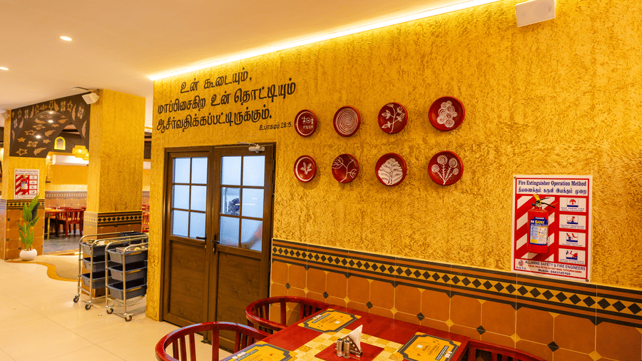 A dining area at DSF Grand Plaza, Tuticorin with a textured gold wall bearing Tamil script, 8 decorative red plates, a wooden door, and a red dining table.