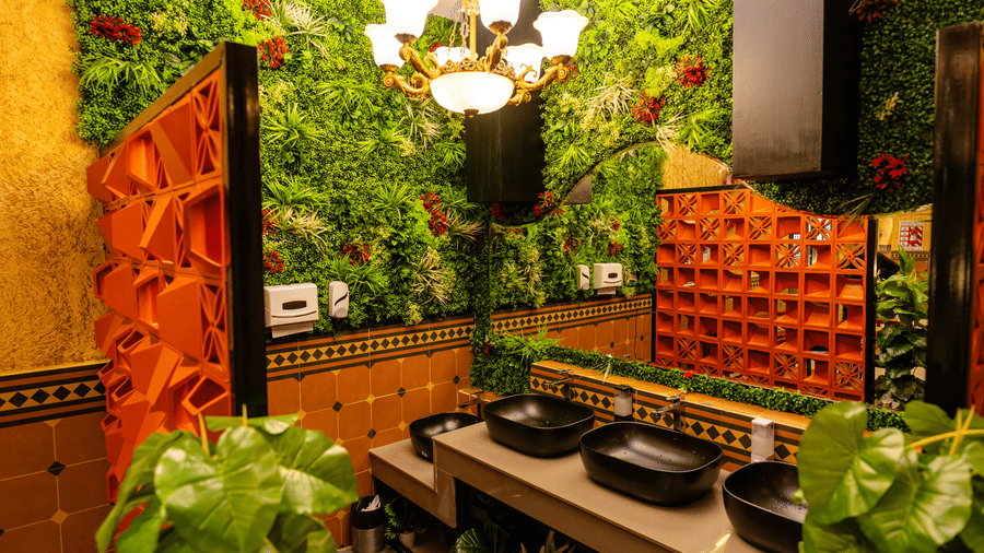A restroom at DSF Grand Plaza, Tuticorin with a green living wall, an ornate chandelier, black oval basins, and an orange geometric partition screen.