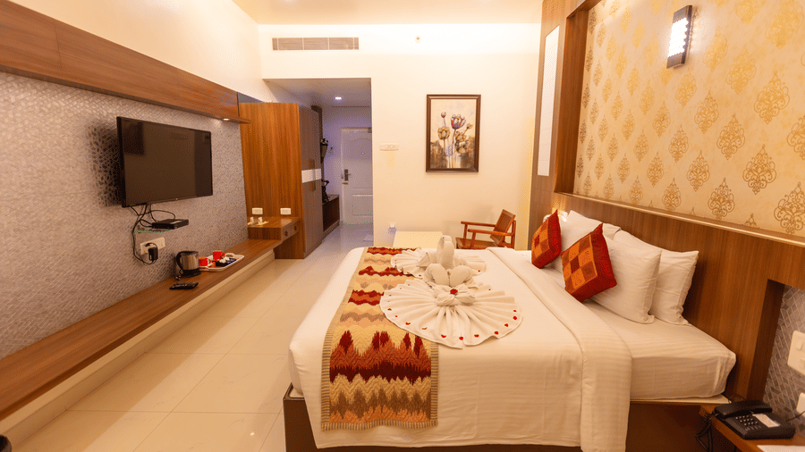 A Royal Executive Room at DSF Grand Plaza, Tuticorin with a king bed dressed with a towel swan, damask wallpaper, a wall-mounted TV, and a wooden rocking chair.