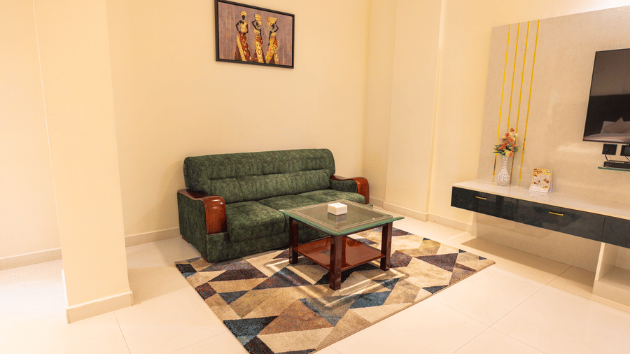 Suite living area at DSF Grand Plaza, Tuticorin with a green sofa, glass-top coffee table, a geometric rug, framed wall art, and a wall-mounted TV.