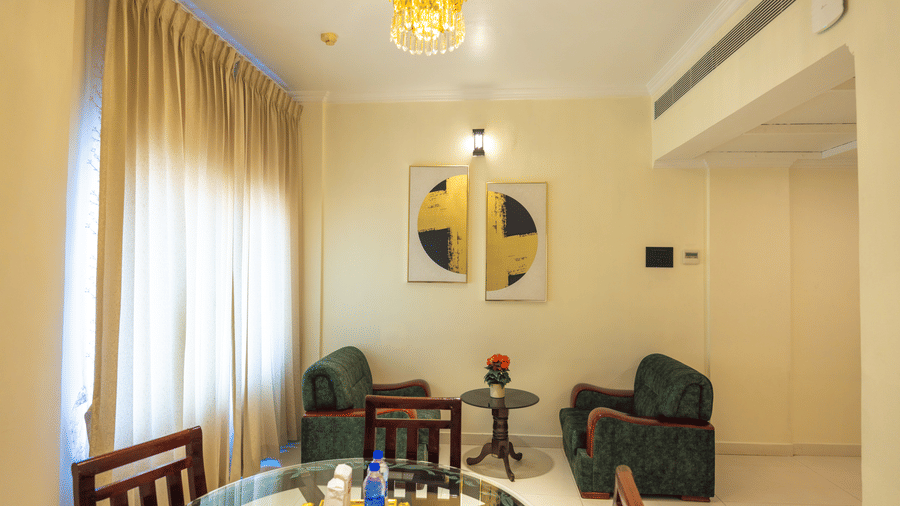 Suite living area at DSF Grand Plaza, Tuticorin with a crystal chandelier, glass dining table, 2 green armchairs, a side table, and framed wall art.