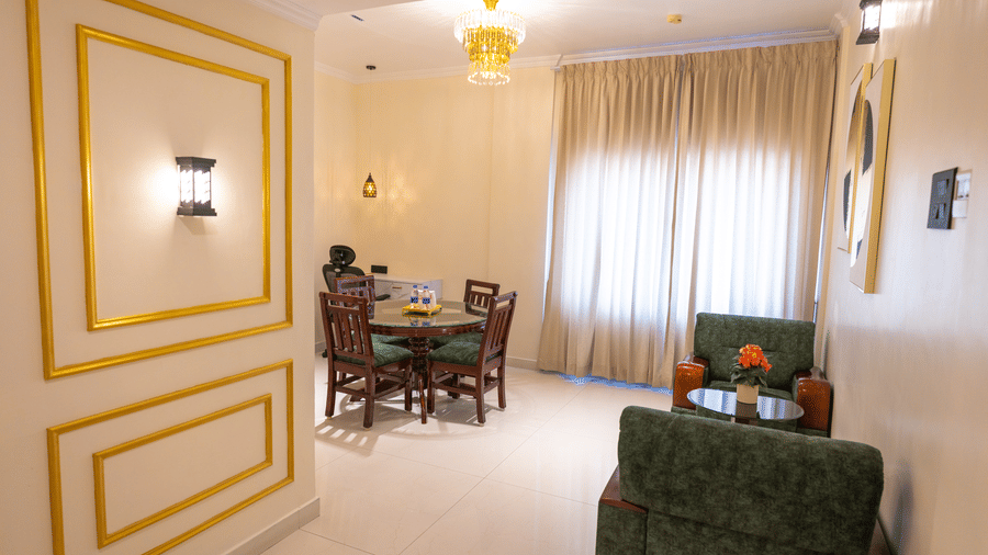 Suite entryway at DSF Grand Plaza, Tuticorin with a gold-framed wall panel, a crystal chandelier, glass dining table, and green armchairs beyond.